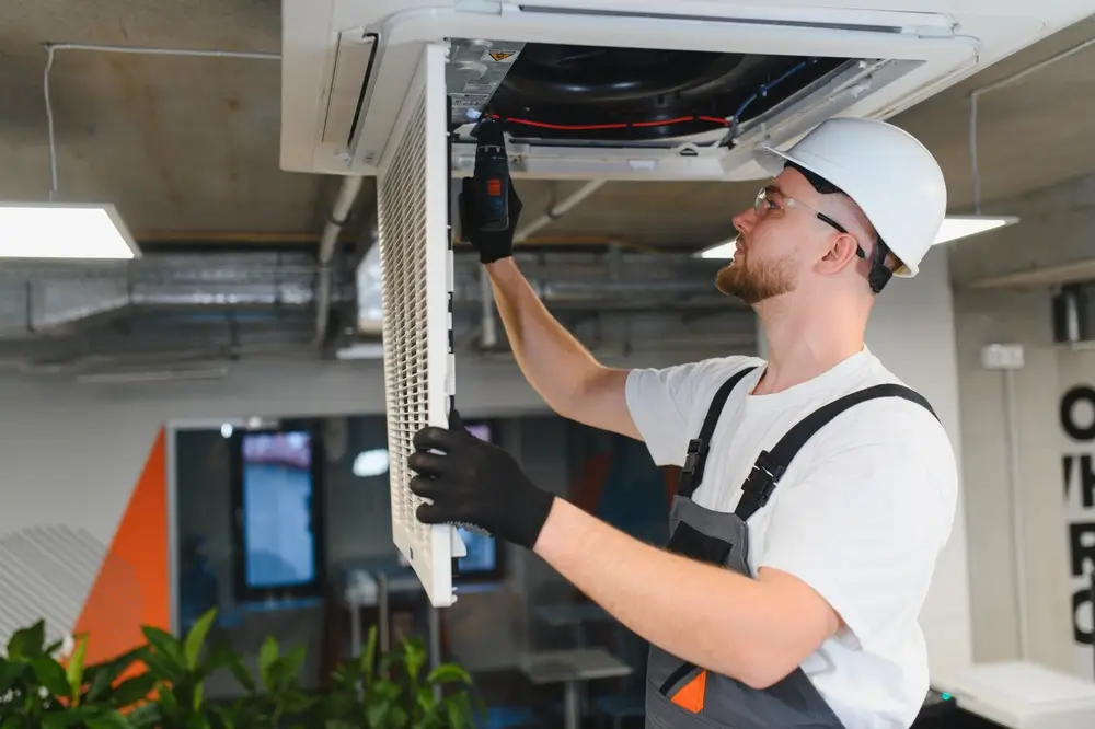 HVAC technician in white hard hat and overalls using a power drill to install a ceiling cassette air conditioning unit inside a commercial building
